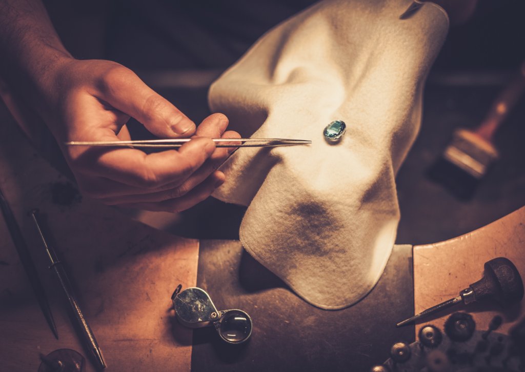 Top-down view of a jeweler with gem tweezers, a soft cloth, and a gem with various jewelers tools around them.